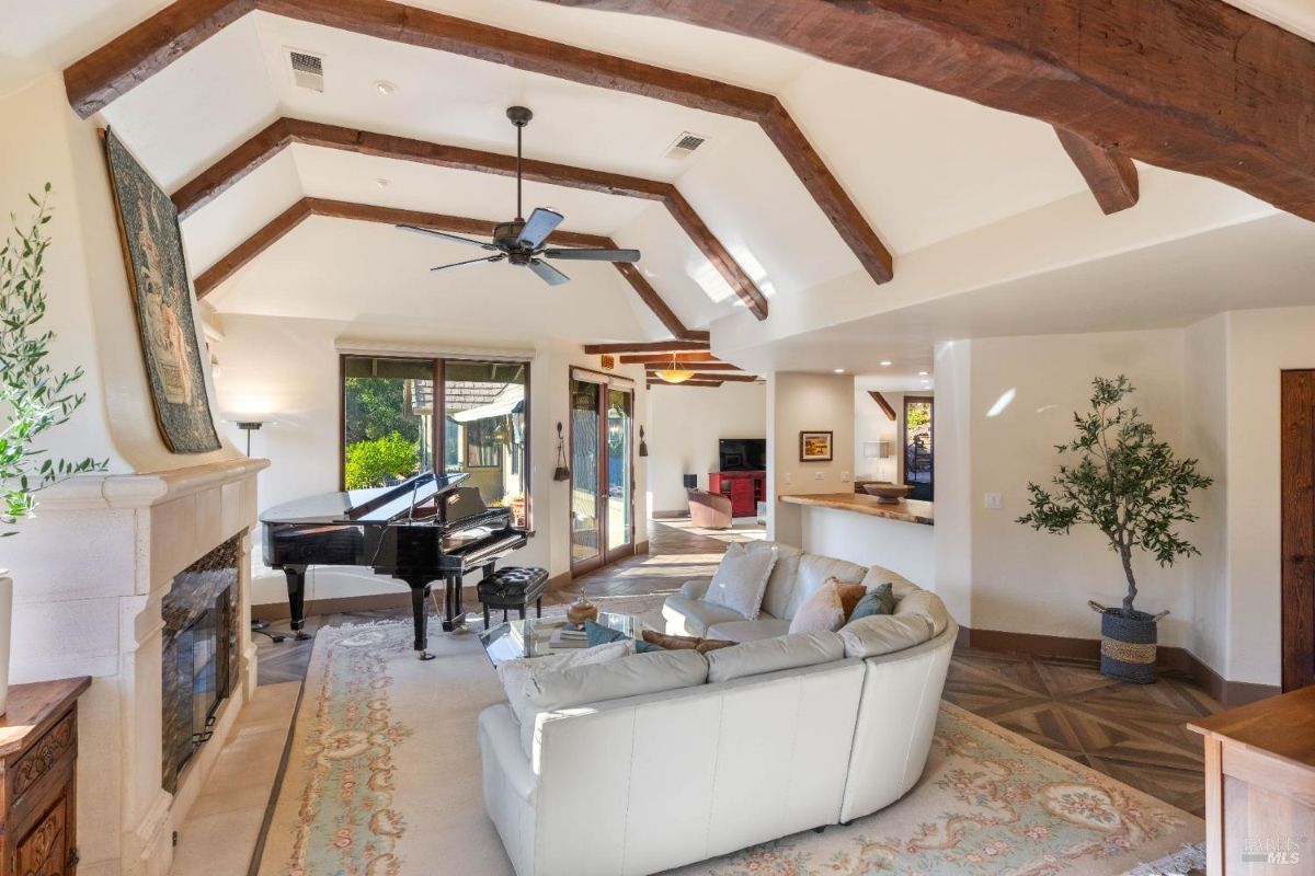 Living area with a fireplace, large windows, and a piano under wooden beam details.