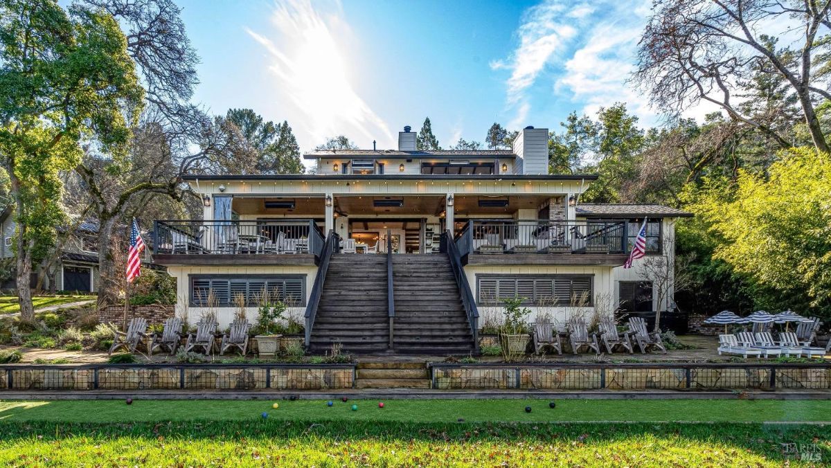 This stately home, with its tiered decks and grand staircase leading down to a lush lawn, exudes an air of relaxed elegance. The American flags fluttering proudly and the bocce ball court add a touch of playful patriotism, creating an inviting space for recreation and leisure.