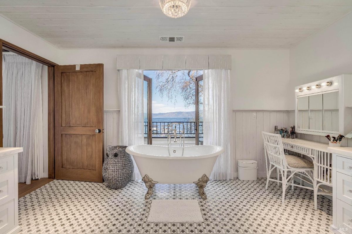 This luxurious bathroom is a haven of relaxation, featuring a freestanding clawfoot tub perfectly positioned to enjoy the stunning waterfront views through the French doors. The elegant chandelier, vanity area, and patterned floor tiles add a touch of glamour, while the white walls and abundant natural light create a sense of spaciousness and serenity.