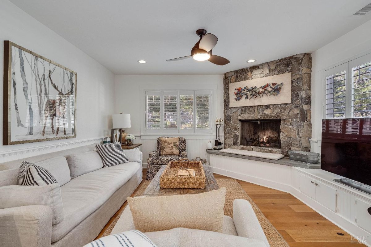 This cozy living room features a charming corner fireplace with a rustic stone surround, creating a warm and inviting focal point. The comfortable seating arrangement, accented by soft furnishings and natural light, encourages relaxation and intimate gatherings.