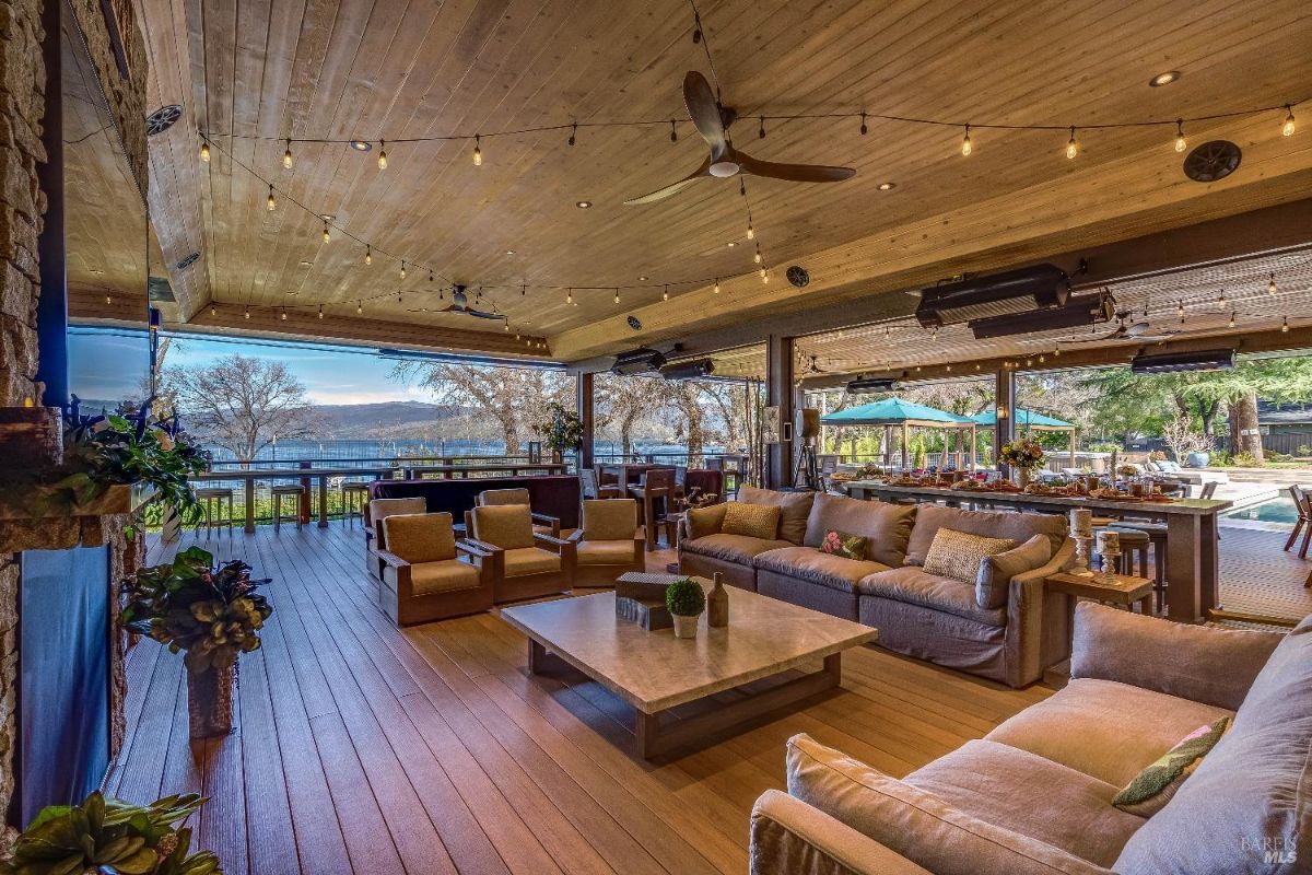 This luxurious outdoor living space is an entertainer's paradise, featuring comfortable seating areas, a cozy fireplace, and a fully equipped bar, all under the warm glow of string lights. The expansive deck overlooks a stunning waterfront view, creating an idyllic setting for relaxation and socializing.