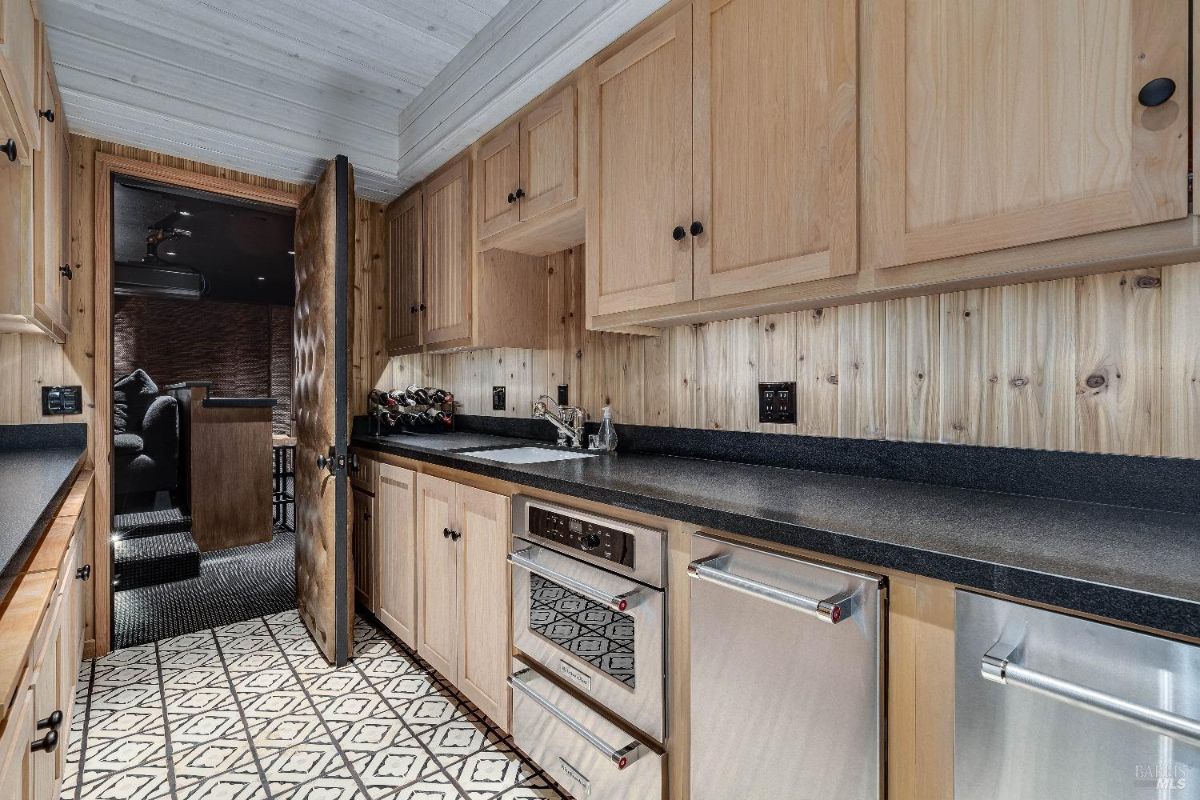 The space is equipped with ample counter space, a sink, an oven, and a dishwasher, making it perfect for preparing snacks and drinks. The wood cabinetry and paneling create a warm and inviting atmosphere, while the patterned tile floor adds a touch of visual interest.