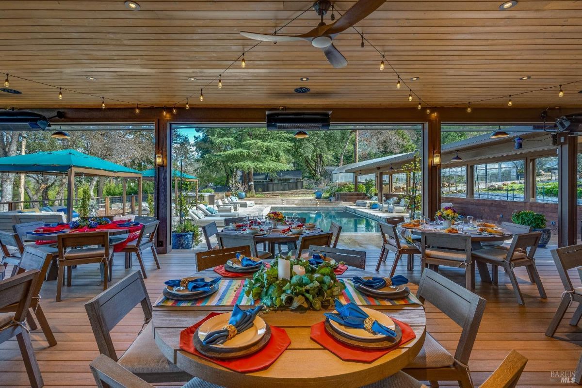 This spacious and festive outdoor dining area is perfect for hosting large gatherings. Multiple tables are set for a feast under a covered patio with string lights, while a sparkling pool and lush greenery create a vibrant backdrop. The open-air design allows guests to enjoy the surrounding landscape and creates a relaxed and inviting atmosphere.