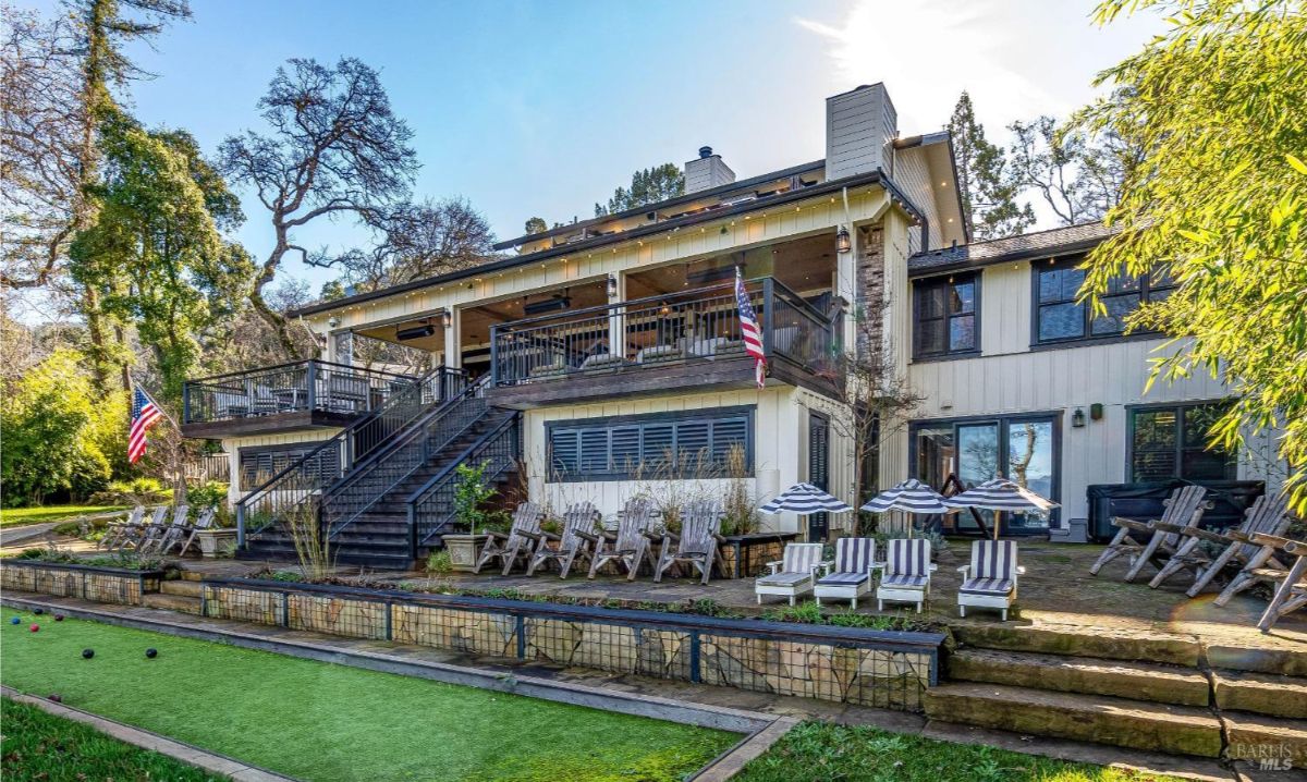 This grand home is an entertainer's dream, boasting expansive tiered decks overlooking a meticulously landscaped yard complete with a bocce ball court. The numerous seating areas and striped umbrellas suggest a relaxed and inviting atmosphere perfect for hosting gatherings and enjoying outdoor leisure.