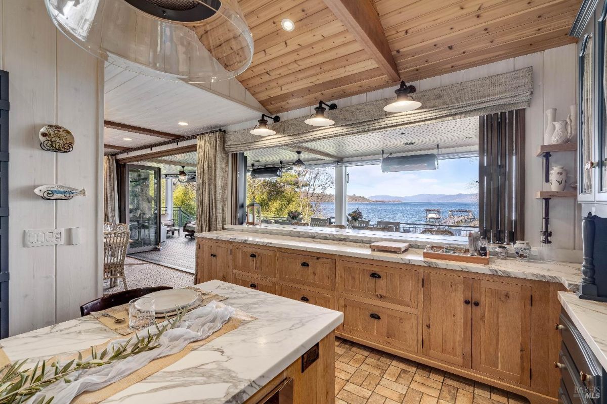 Kitchen features a long countertop with ample workspace and cabinetry, perfect for meal preparation. The large window above the counter offers stunning views of a waterfront scene, creating a serene and inspiring backdrop for culinary creations.