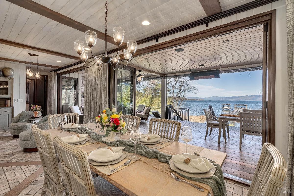 This elegant dining room seamlessly blends indoor and outdoor living, with expansive glass doors that open onto a waterfront deck. The rustic dining table, set for a feast, is bathed in natural light, creating an inviting space for gatherings and celebrations.