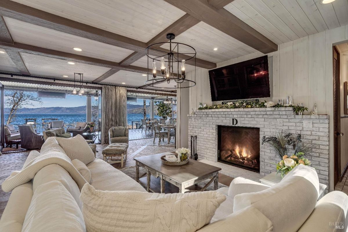 This luxurious living room is designed for both comfort and entertainment. A roaring fire in the white brick fireplace adds warmth, while the expansive windows offer breathtaking views of a waterfront landscape, seamlessly blending indoor and outdoor living.