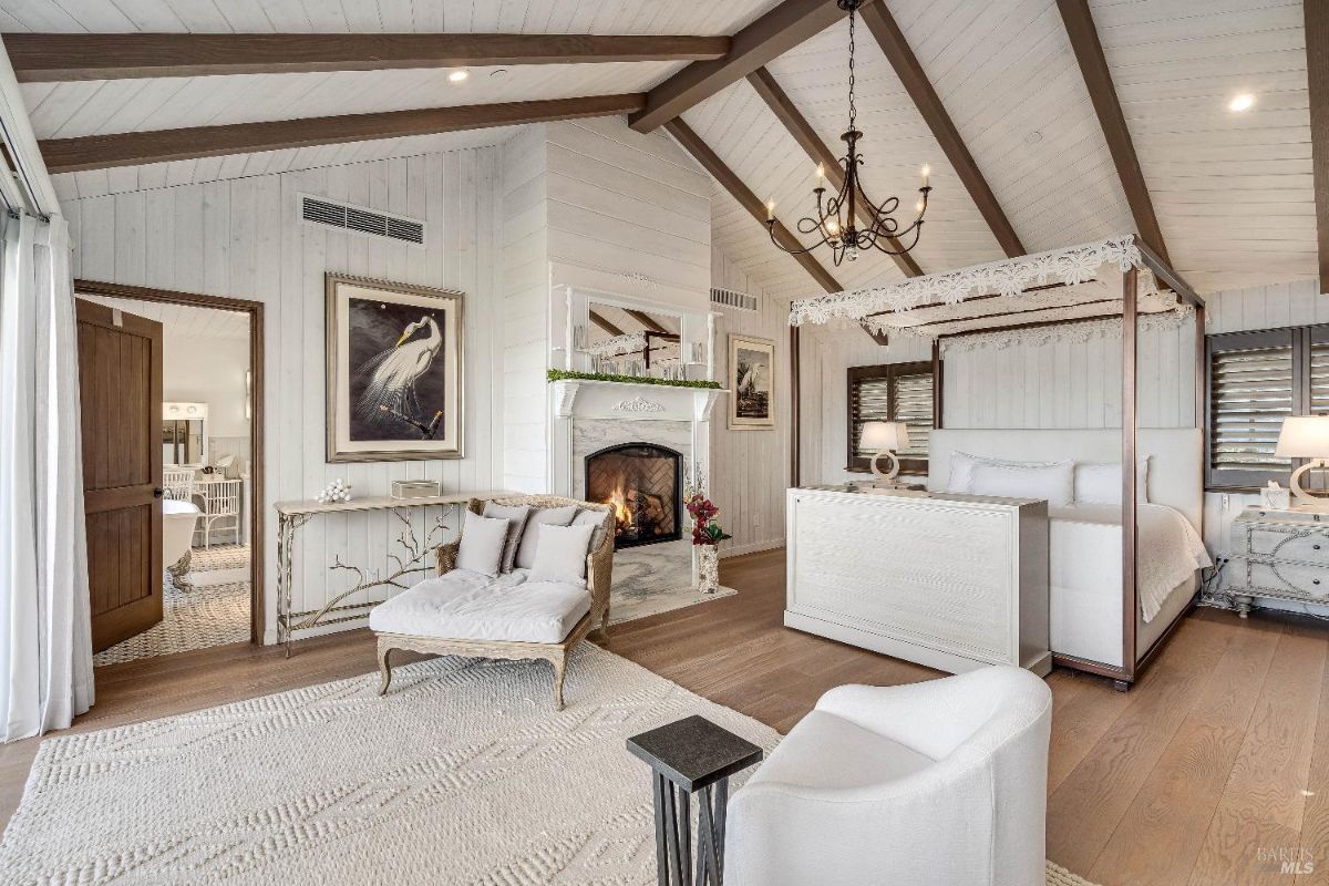 This luxurious bedroom exudes a serene and romantic ambiance, with a cozy fireplace, a charming chaise lounge, and a beautiful canopy bed. The high ceilings with exposed beams and the plush furnishings create a sense of grandeur and comfort, while the artwork and décor add a touch of elegance.