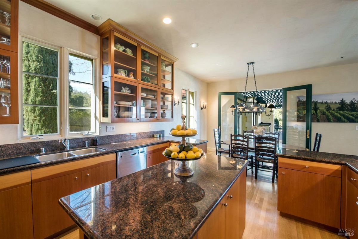 A spacious kitchen with wooden cabinets, granite countertops, and stainless steel appliances. A large island with a breakfast bar sits in the center of the room, and a dining table with chairs is visible in the background.