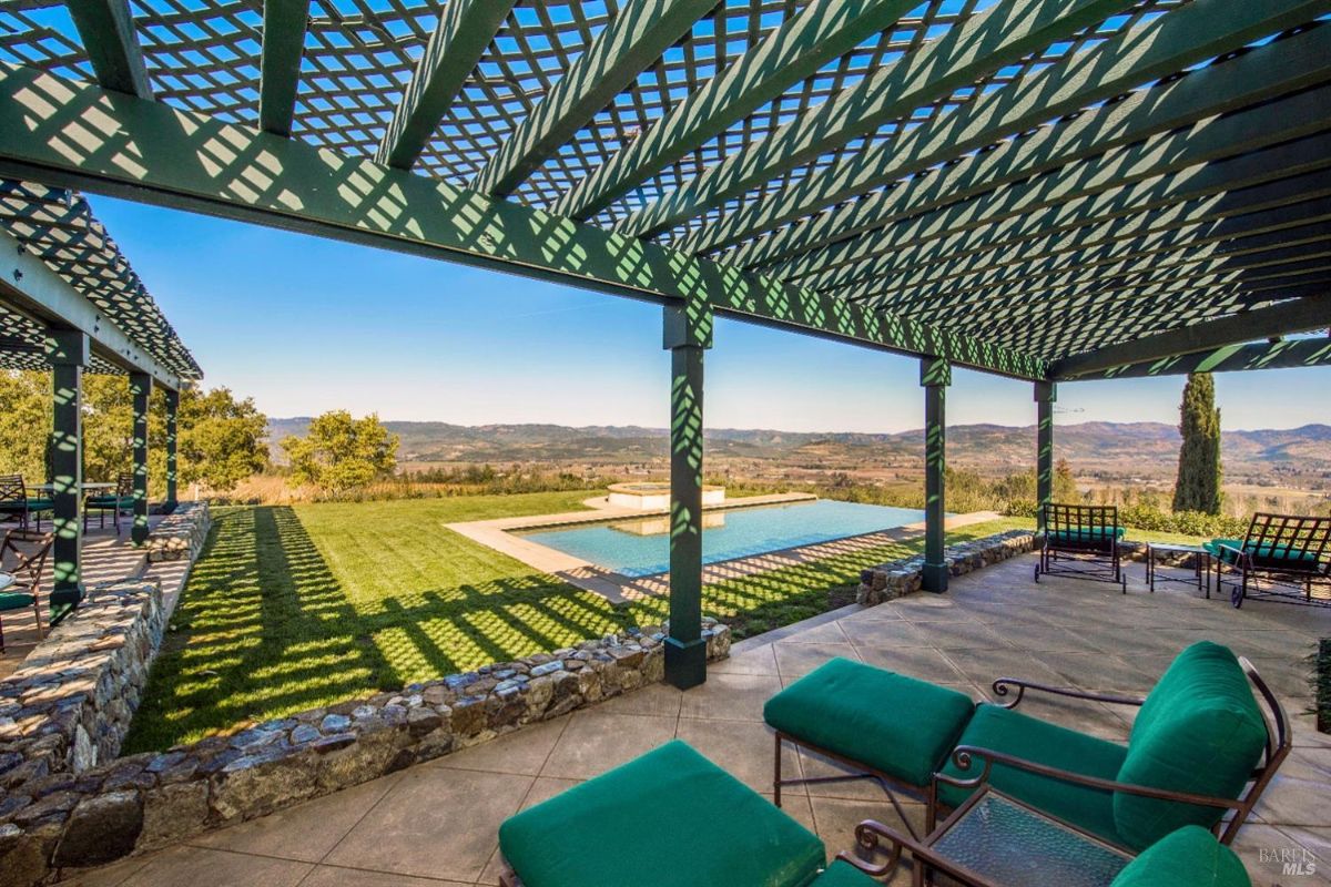 A covered patio with a swimming pool in the background. The patio has a wooden lattice roof and is furnished with comfortable chairs and loungers. The pool is surrounded by a grassy area and offers stunning views of rolling hills in the distance.