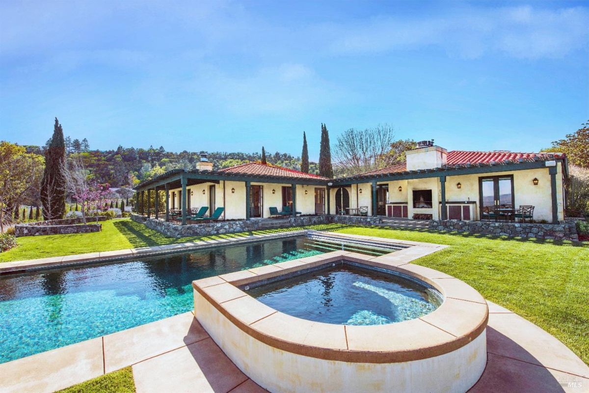 A large house with a swimming pool and a hot tub in the foreground. The house has a tiled roof, a stone facade, and a covered patio. The sky is blue and there are trees and hills in the background.