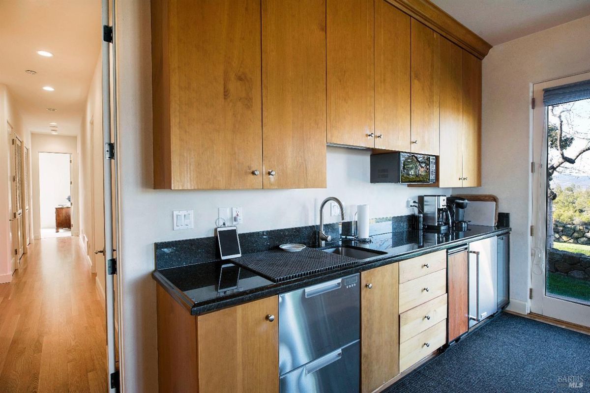 A small, well-equipped kitchen with wooden cabinets, granite countertops, and stainless steel appliances. There's a sink, a microwave, a coffee maker, and a small refrigerator. A door leads to a hallway, and a window offers a view of the outdoors.