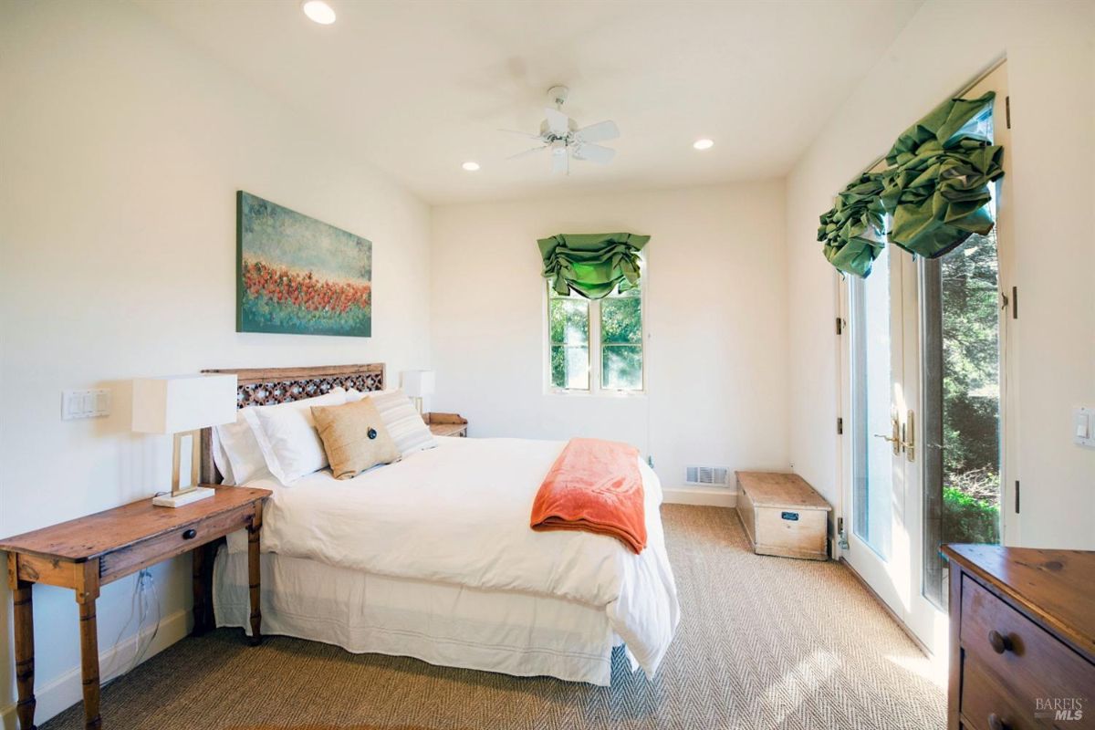 A bright and airy bedroom with a king-sized bed, a large painting on the wall, and a pair of doors leading to a balcony. The room has a window with green curtains and a bedside table with a lamp.