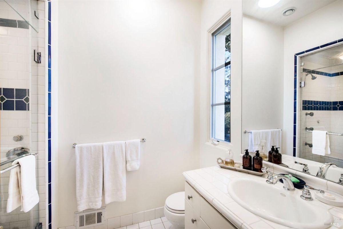 Bright and clean bathroom with white tiles and blue accents. There is a single sink, a toilet, and a shower stall with glass doors. A window provides natural light and a view of the outdoors.