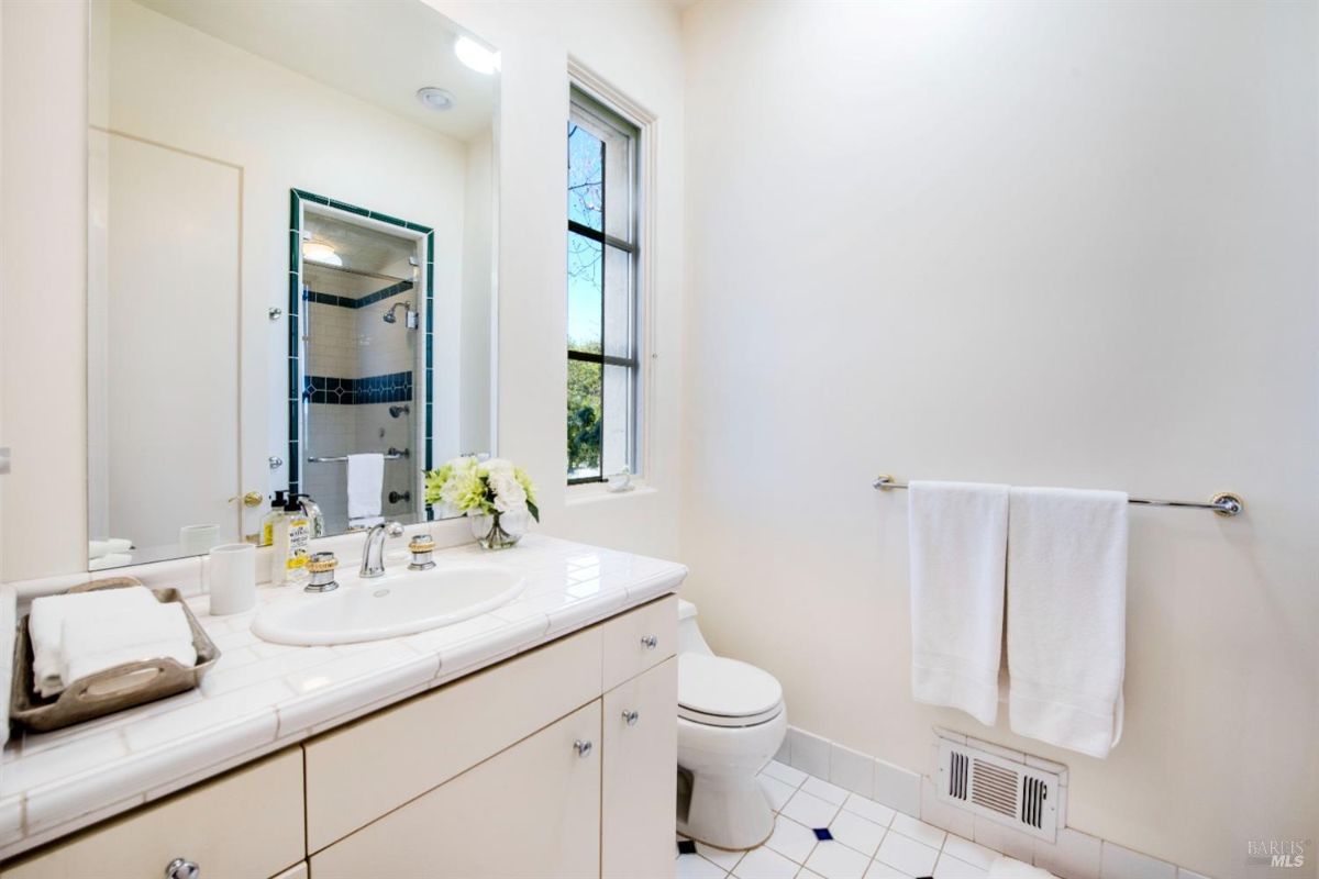 A clean, white bathroom with a single sink, a toilet, and a shower stall visible in the background. There is a towel rack with two white towels and a window with a view of trees.