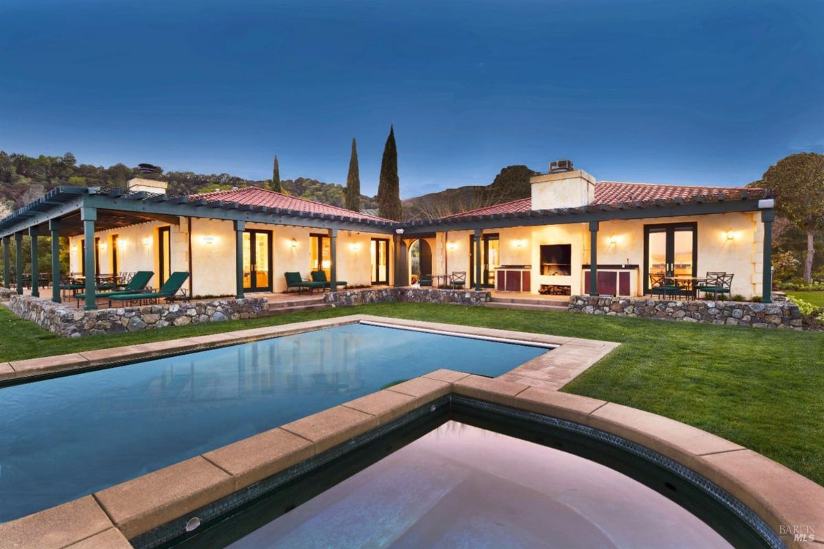 Large house with a swimming pool in the foreground. The house has a tiled roof, a stone facade, and a covered patio. The pool is illuminated, and the sky is a deep blue.