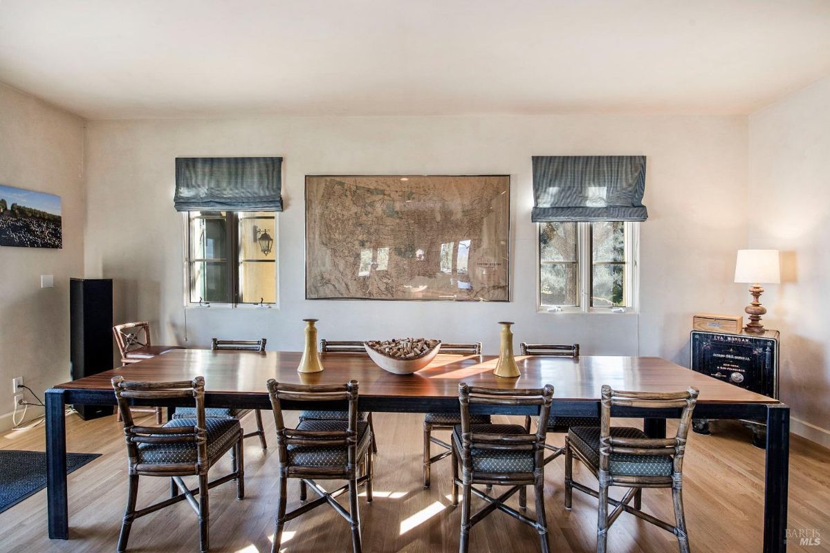 A spacious dining room with a long wooden table, eight chairs, and a large framed map hanging on the wall. There are two windows with curtains on either side of the map.