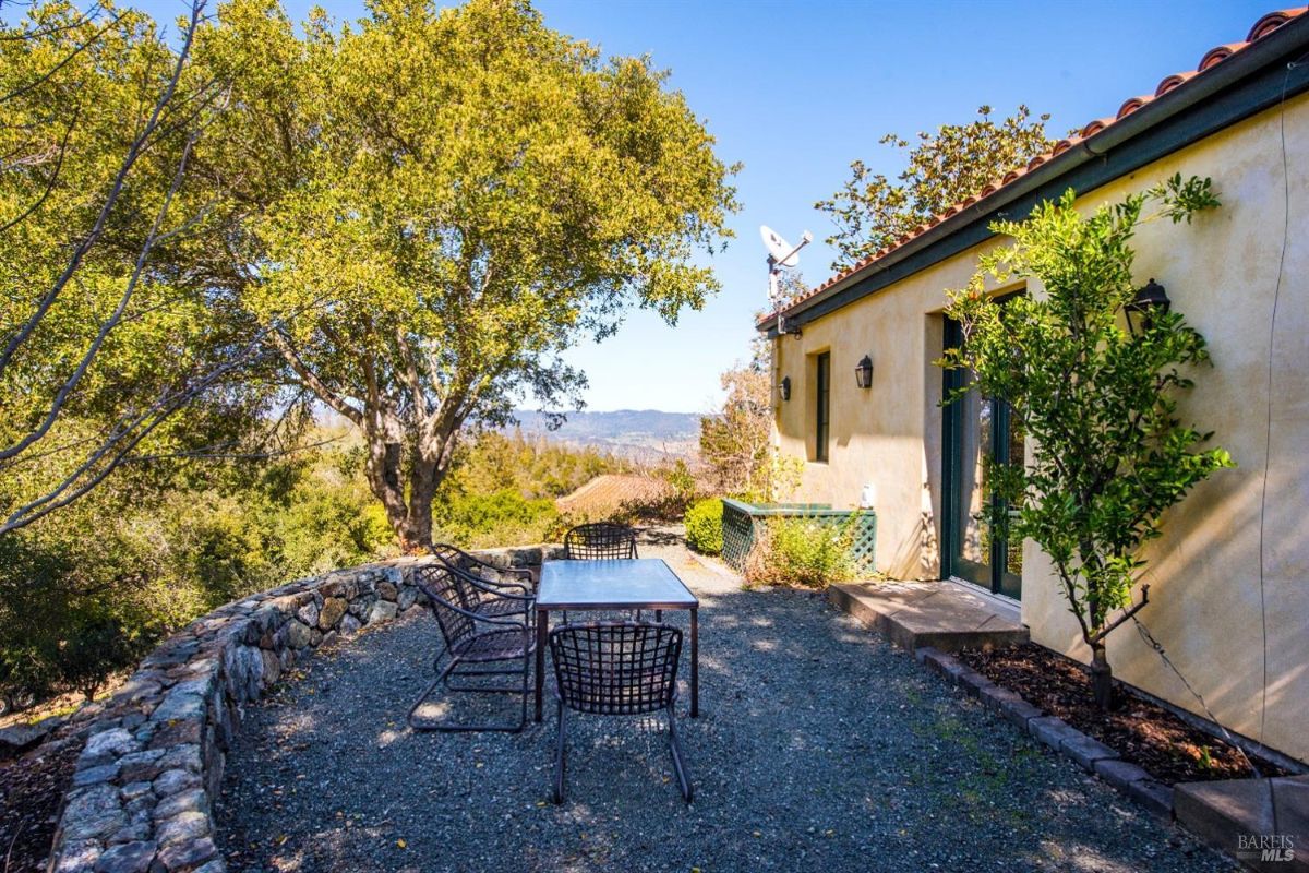 Small patio with a table and chairs. Located next to a house with a red tile roof. Overlooks a scenic view of rolling hills. Surrounded by mature trees and a stone wall.