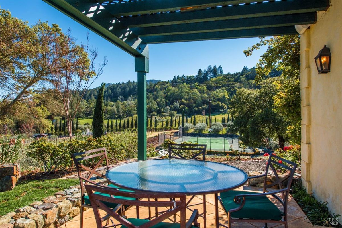 Covered patio with wooden lattice roof and green pillars. Overlooks a lush green lawn, tall trees, and a vineyard. Tennis court visible in the background.