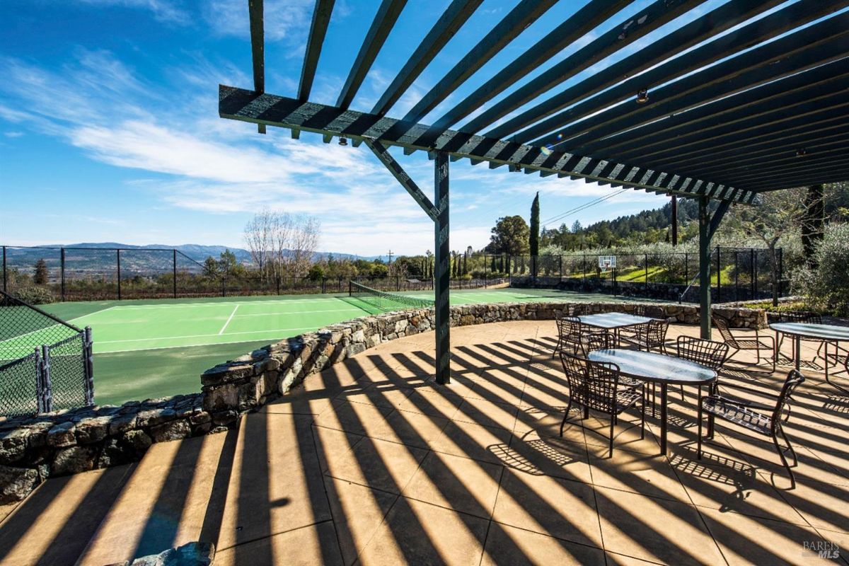 A covered patio with a wooden lattice roof overlooks a tennis court. The patio has several tables and chairs, and there are beautiful views of rolling hills in the distance.