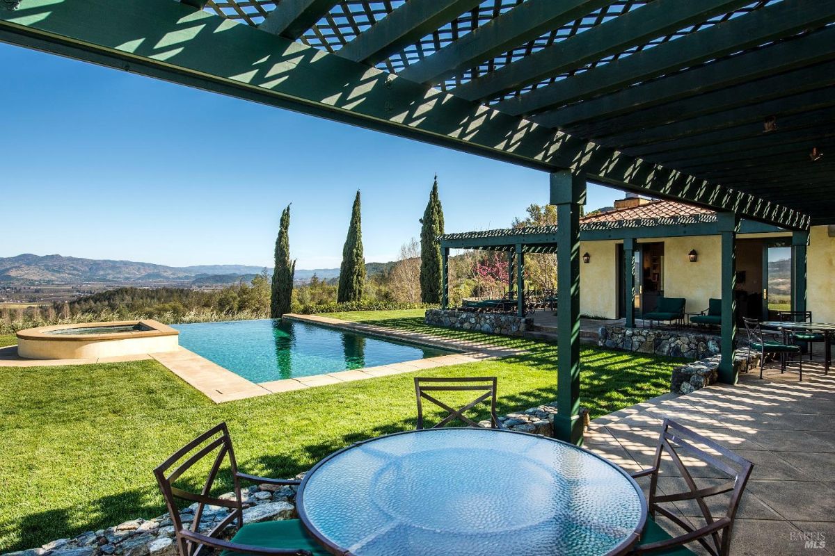 A covered patio with a wooden lattice roof overlooks a swimming pool and a hot tub. The patio has a table and chairs, and there are beautiful views of rolling hills in the distance.