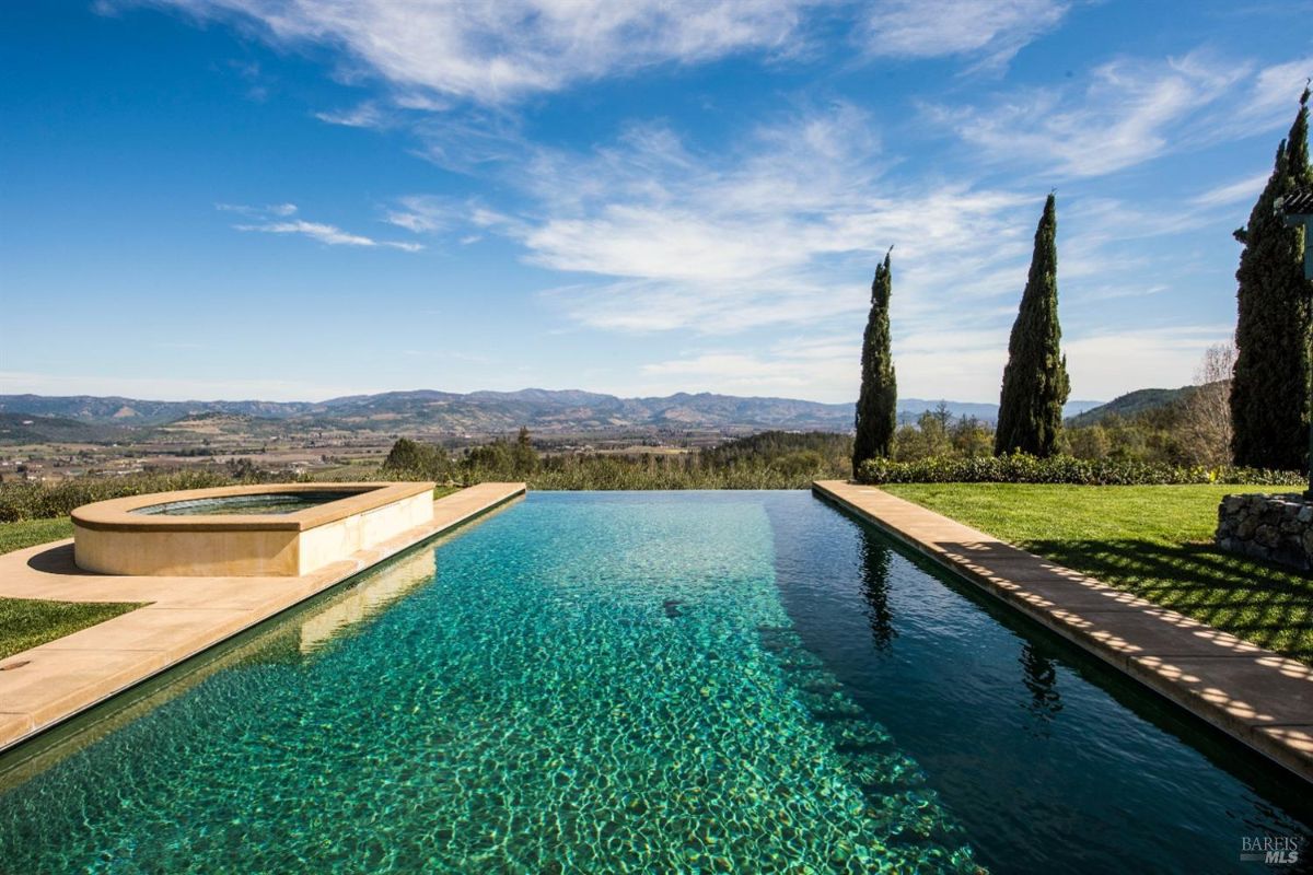 Large infinity pool with a hot tub overlooking a scenic valley. Blue sky with white clouds.