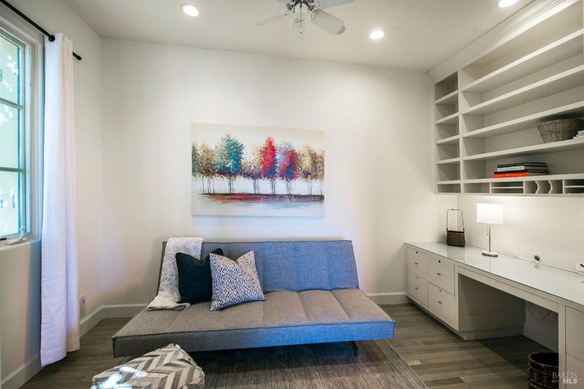 A bright, modern office space with a blue sofa, a desk with a glass top, and built-in white shelves. A large colorful painting hangs on the wall above the sofa.