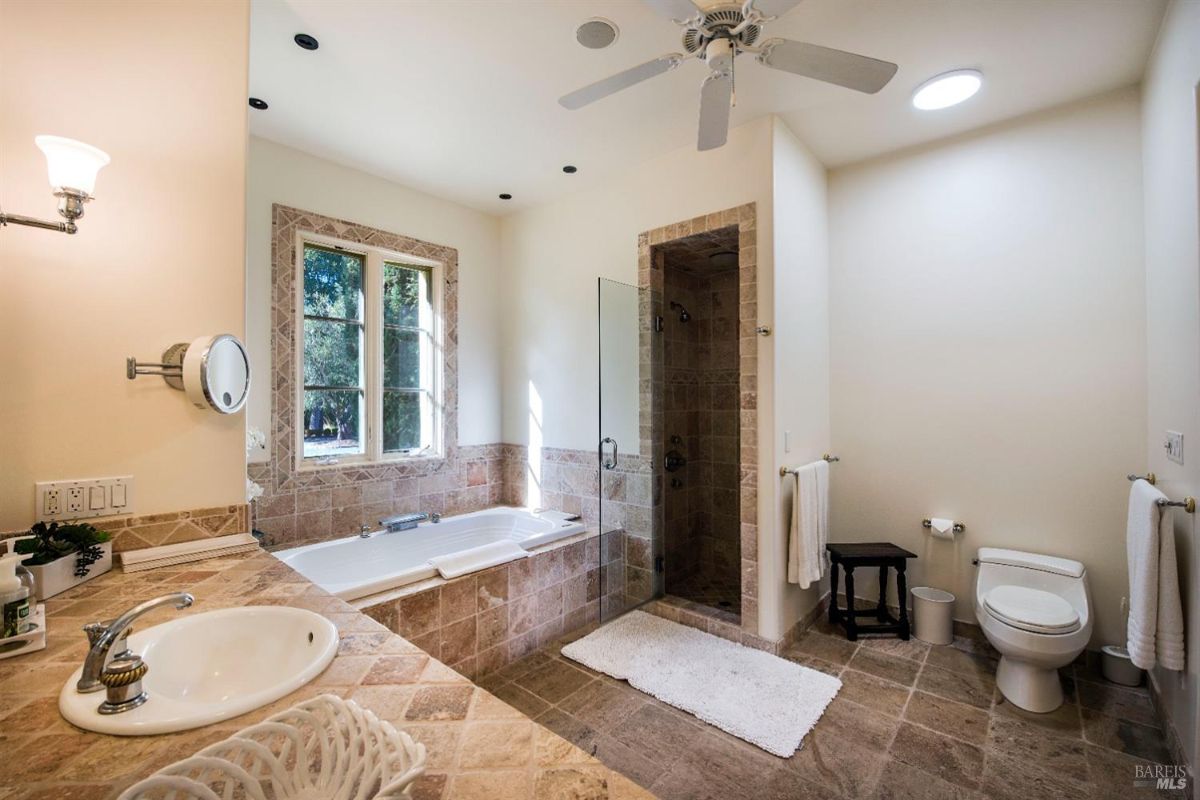 Bathroom with a large bathtub, a separate shower, and a double vanity. The room has a window with a view of the outdoors and a ceiling fan.