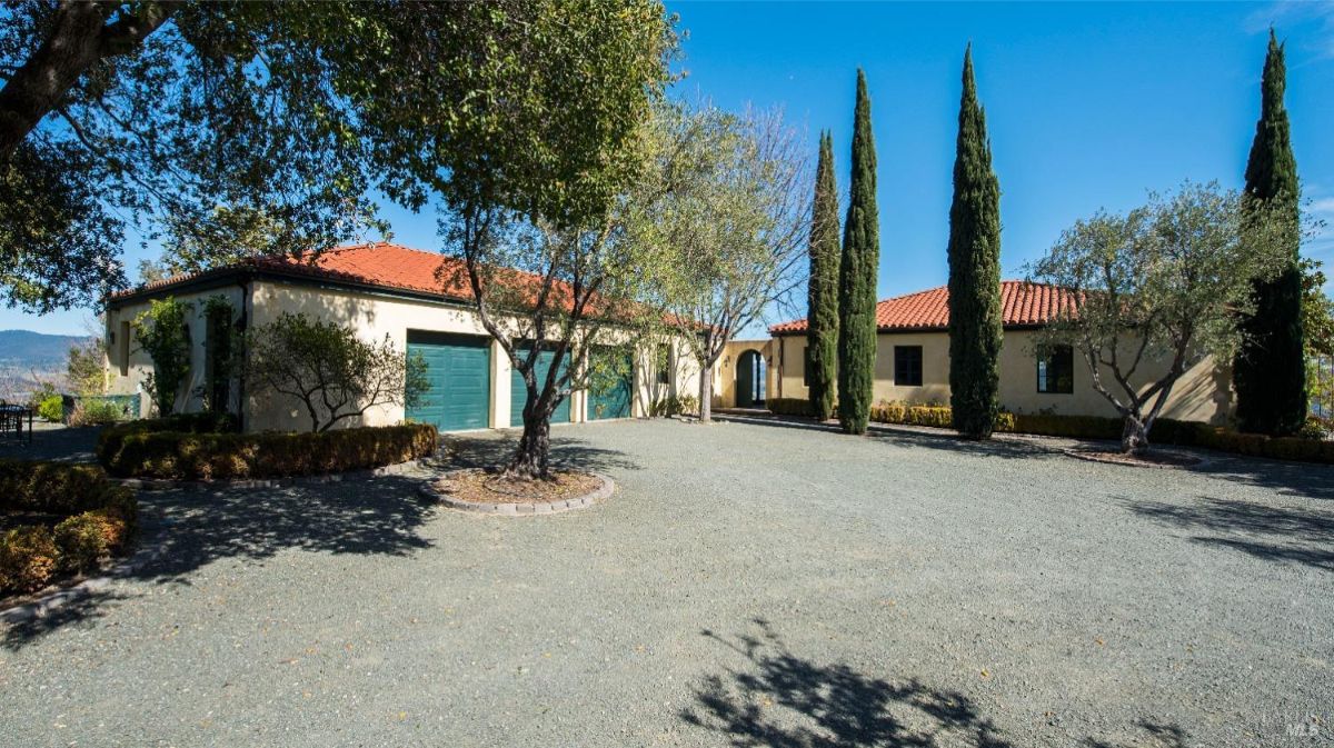 Large house with a red tile roof and a garage. Surrounded by a gravel driveway and mature trees. Located on a hilltop with a view of the surrounding landscape. The house has a classic Mediterranean style architecture.