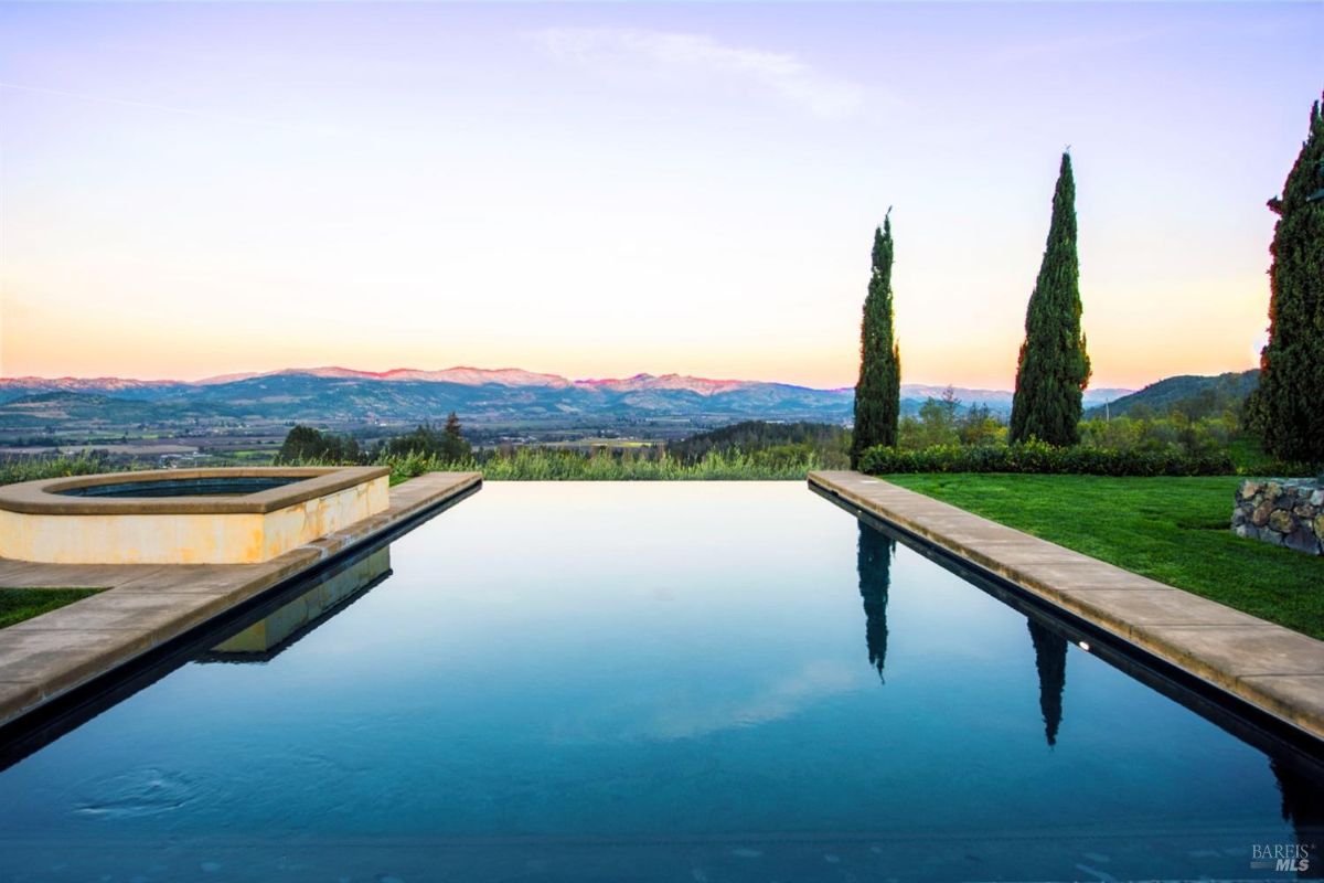 Large infinity pool with a hot tub. Overlooks a scenic valley. Surrounded by a grassy area and tall trees. The pool reflects the beautiful sunset sky.