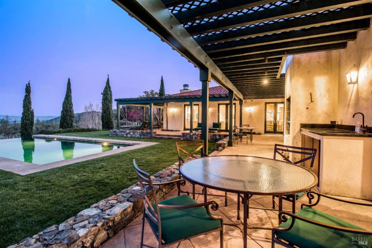 A covered patio with a swimming pool in the background. The patio has a wooden lattice roof and is furnished with a round table and chairs. The pool is surrounded by a grassy area and offers stunning views of rolling hills in the distance. The sky is twilight, creating a serene atmosphere.