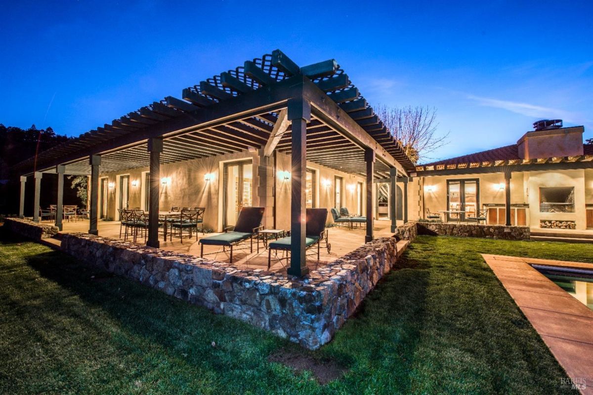 Large house with a covered patio illuminated by warm lights. The patio has a wooden lattice roof and is furnished with comfortable chairs and loungers. The house is surrounded by a well-manicured lawn and a swimming pool can be seen in the background.