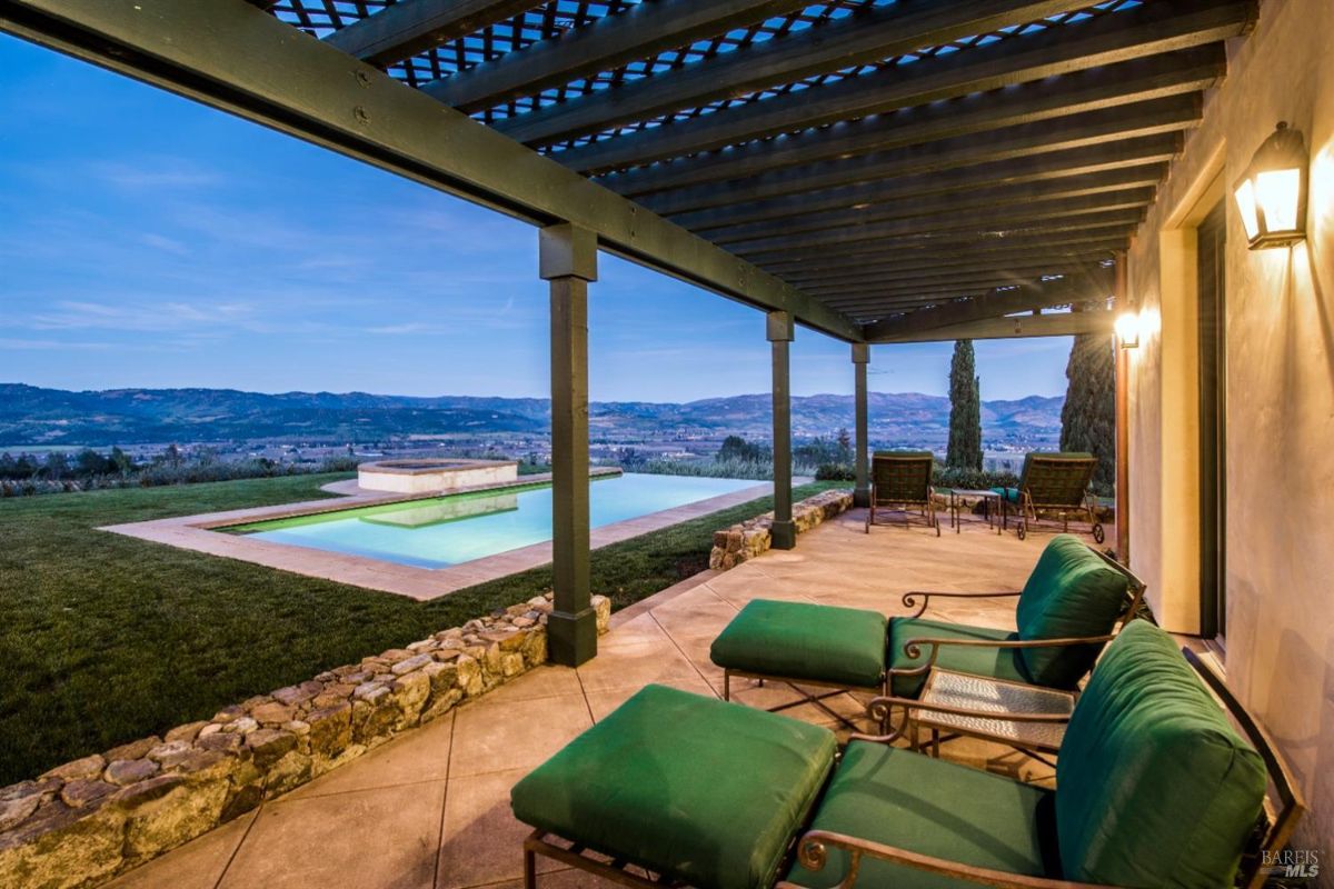 A covered patio with a swimming pool in the background. The patio has a wooden lattice roof and is furnished with comfortable chairs and loungers. The pool is surrounded by a grassy area and offers stunning views of rolling hills in the distance. The sky is twilight, creating a serene atmosphere.