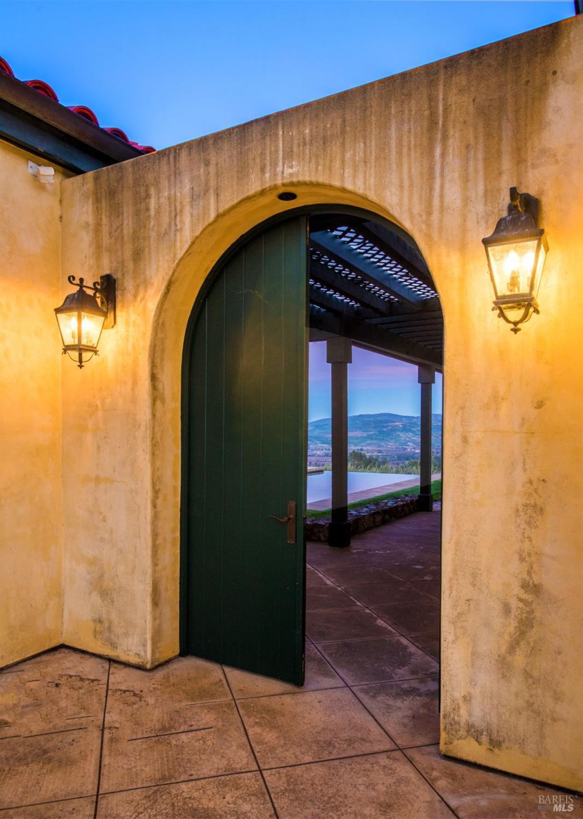 An arched doorway with a dark green door leads to a patio with a view of rolling hills in the distance. The wall is a warm yellow color and there are two wall-mounted lanterns on either side of the door.