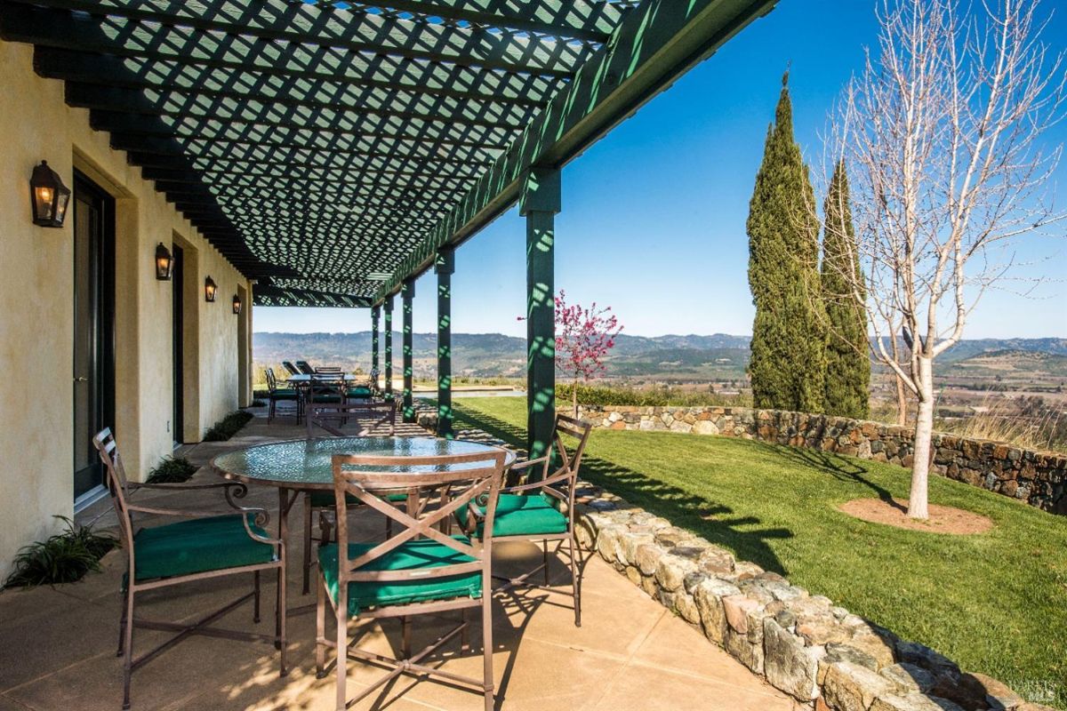 Covered patio with a wooden lattice roof and green pillars. Overlooks a lush green lawn, tall trees, and a vineyard in the distance.