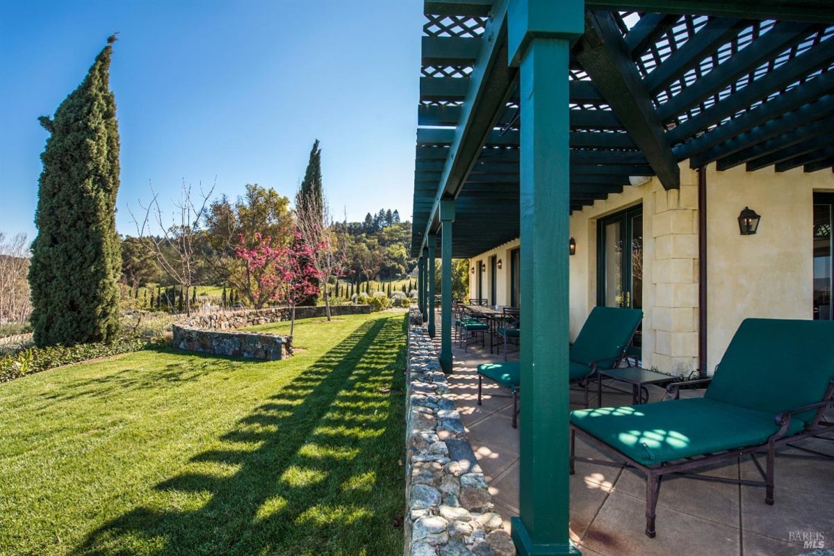 Patio with a wooden lattice roof and green pillars. The patio is furnished with comfortable chairs and loungers. A lush green lawn stretches out in front, and there are tall trees and a vineyard in the background.