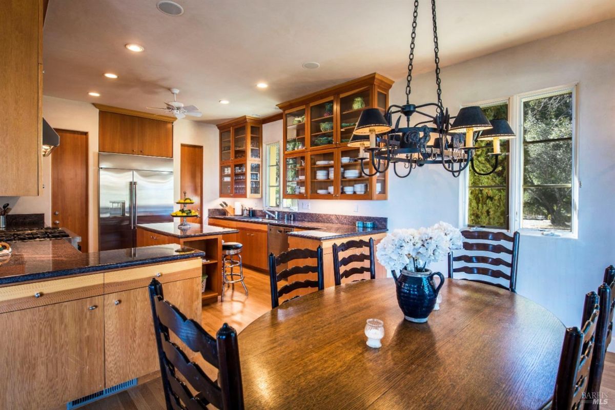 Kitchen with wooden cabinets, granite countertops, and stainless steel appliances. A large dining table with chairs is positioned in the center of the room, and a chandelier hangs above it.