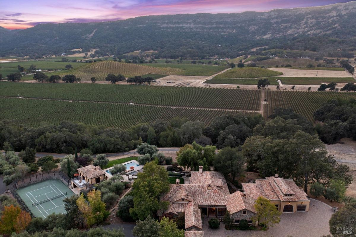 Aerial view of a large estate with a Mediterranean-style mansion, tennis court, swimming pool, and extensive vineyards in the background. The property is situated in a picturesque valley with rolling hills.