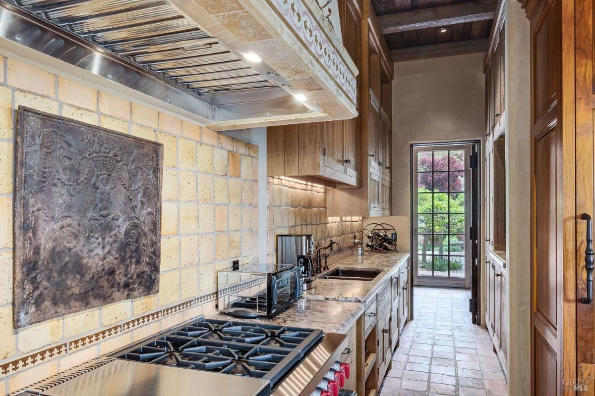 Galley kitchen with wooden cabinetry, tiled backsplash, and a professional-grade gas cooktop. Features a marble countertop and a glass door leading to an outdoor area.