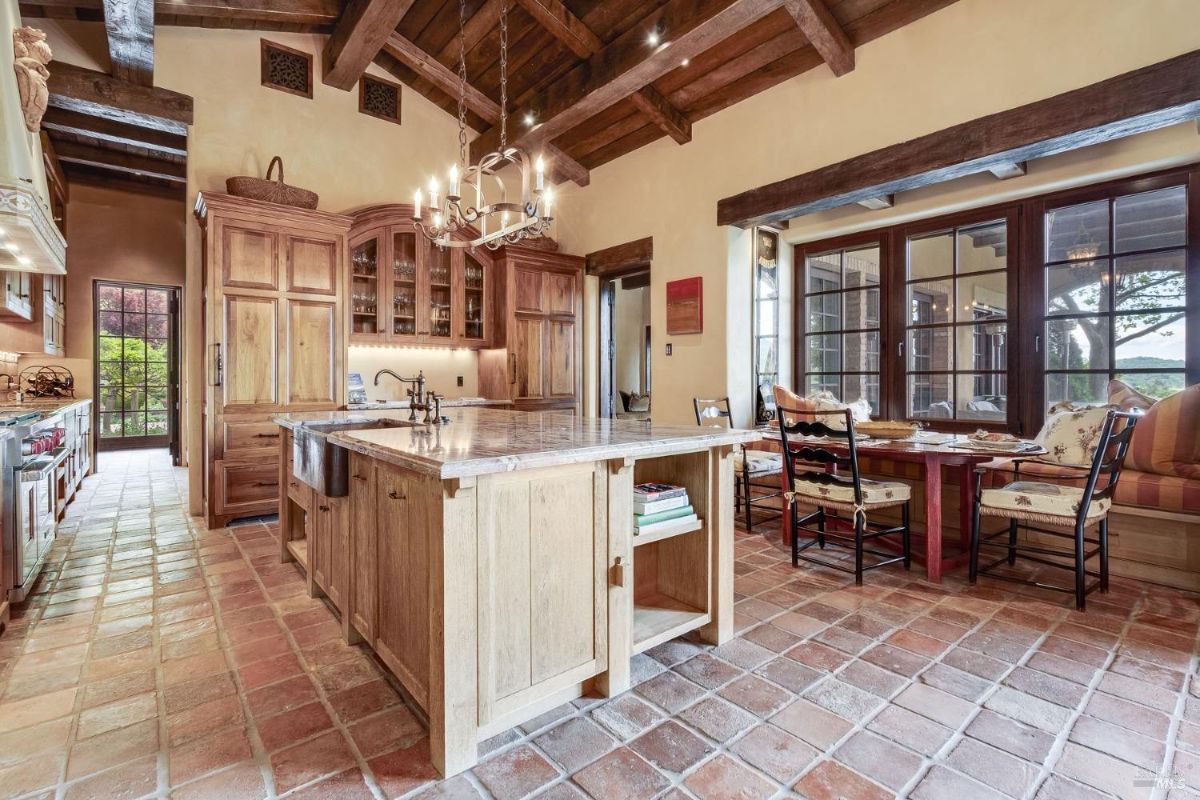 Kitchen with exposed wooden beams, built-in cabinets, and an island featuring a farmhouse-style sink and marble countertop. Adjacent dining area includes built-in bench seating by large windows with views of the outdoors.