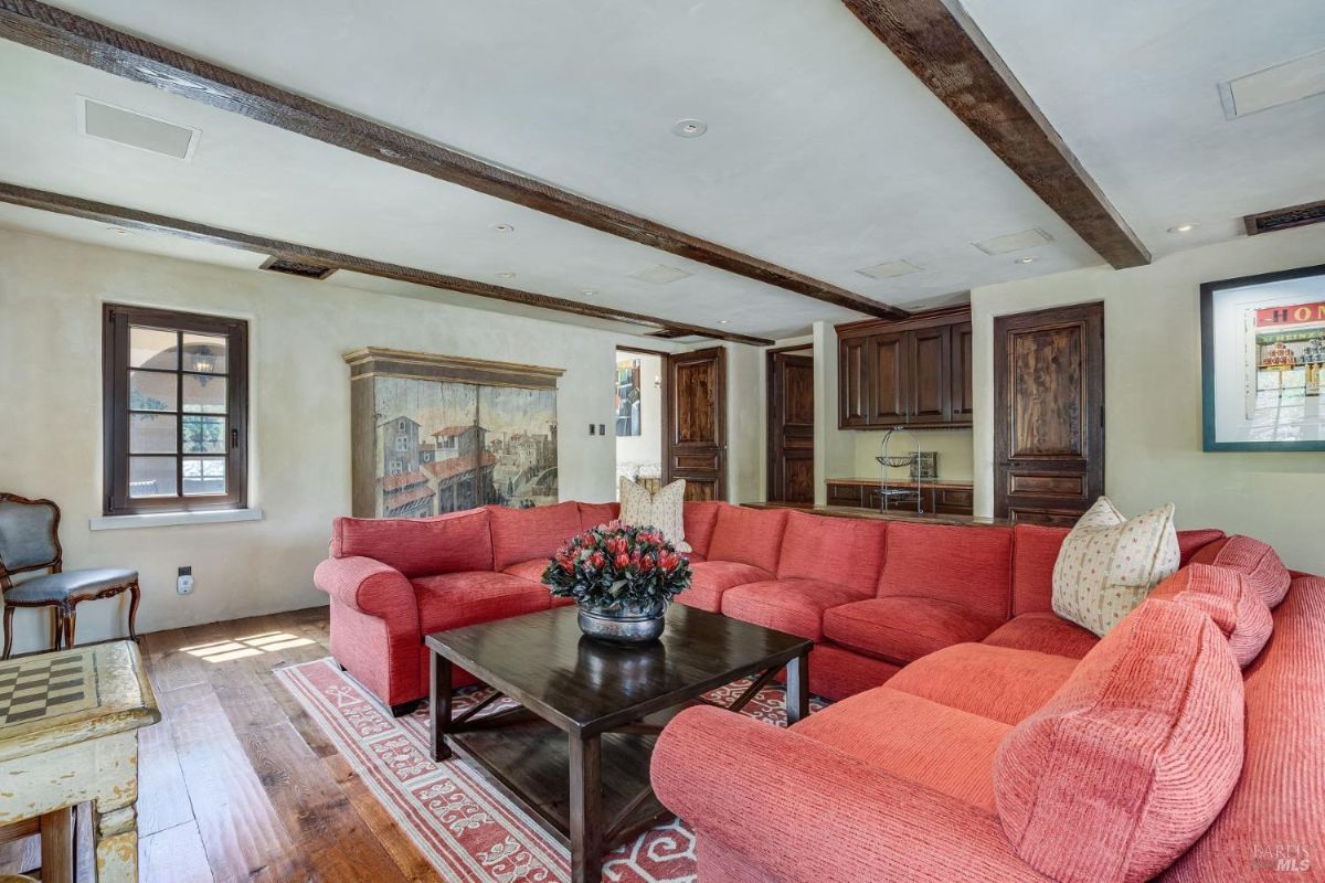 Living room features a red sectional sofa surrounding a dark wood coffee table on a patterned area rug. Wooden ceiling beams, a built-in cabinet, and a painted wall panel add architectural and decorative elements to the space.