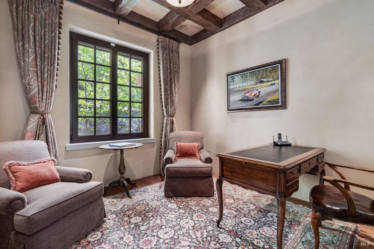 Room features two armchairs with accent pillows, a small side table, and a wooden desk with a leather chair. Large window with patterned curtains provides natural light, and a framed artwork hangs on the wall.