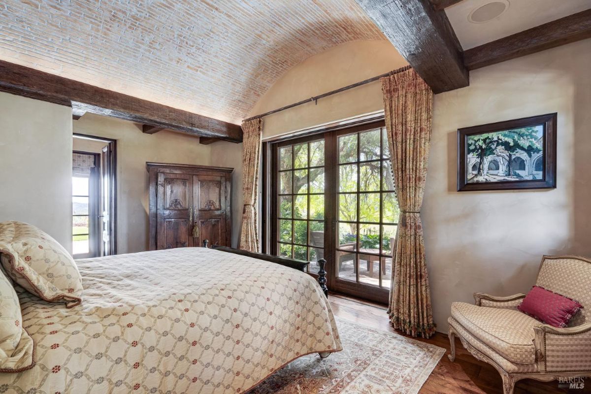 Bedroom with a vaulted brick ceiling, exposed wooden beams, and glass doors opening to a lush outdoor view. Features an antique wardrobe, patterned curtains, and an upholstered armchair.