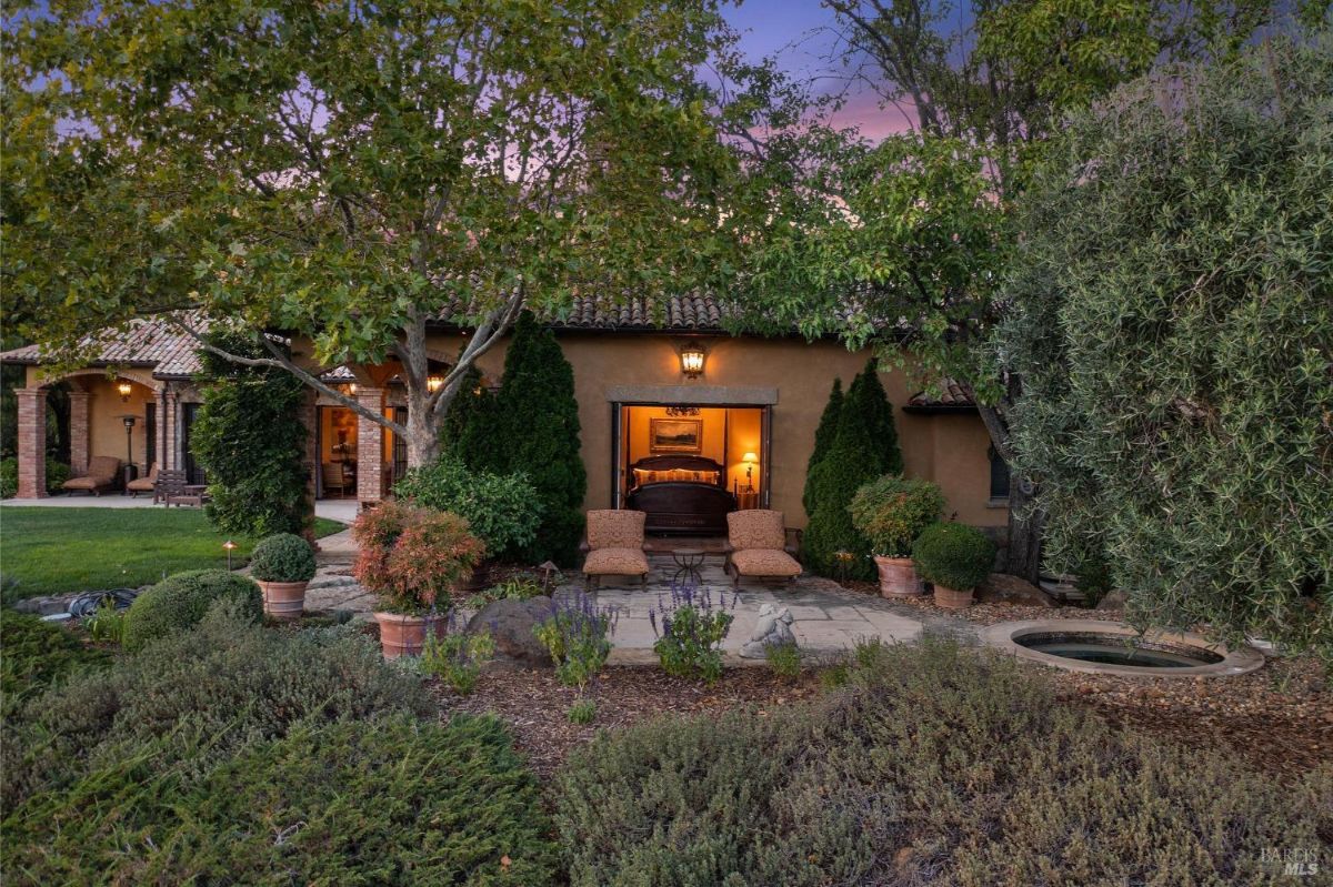 Exterior view of a Mediterranean-style villa with lush greenery and an open patio connected to a cozy bedroom.