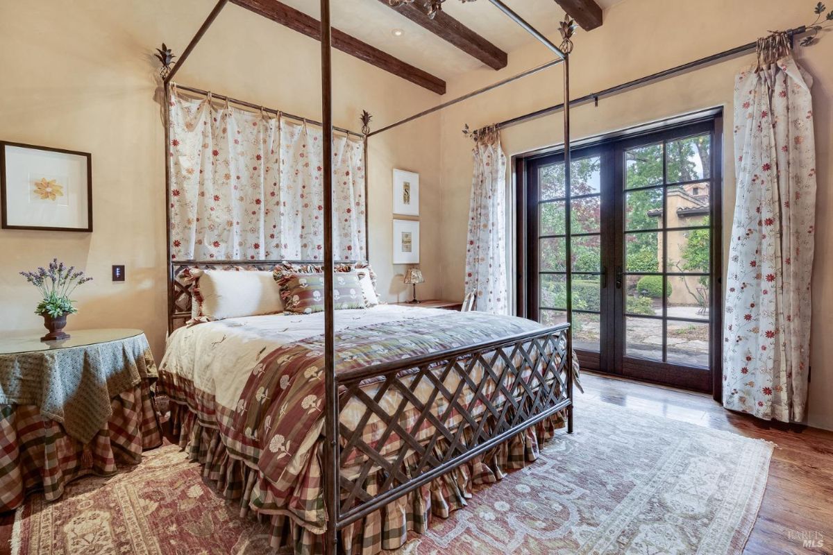 Bedroom with exposed wooden beams and a four-poster bed featuring floral-patterned curtains and bedding. Glass doors open to an outdoor patio, complemented by a vintage area rug and a skirted side table.