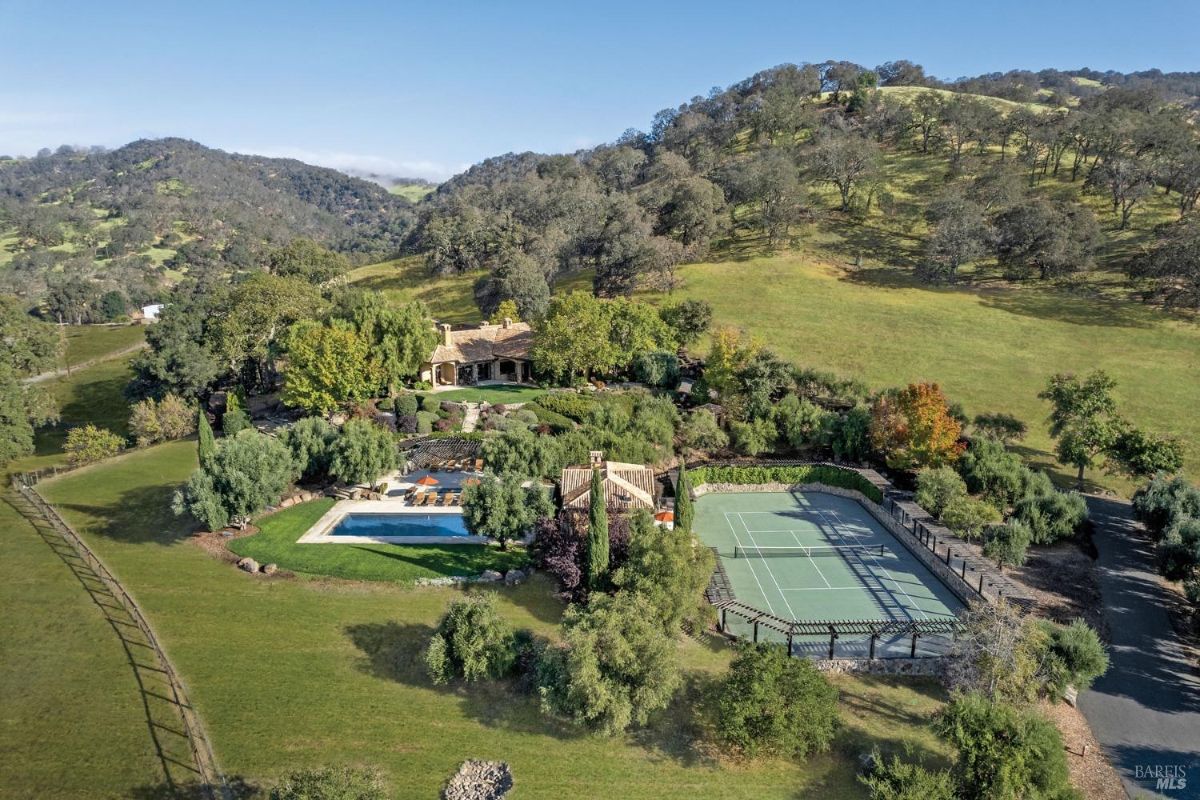 Sprawling estate features a central residence surrounded by landscaped gardens, trees, and hilly terrain. Includes a swimming pool with lounge area and a tennis court enclosed by pergolas and greenery.