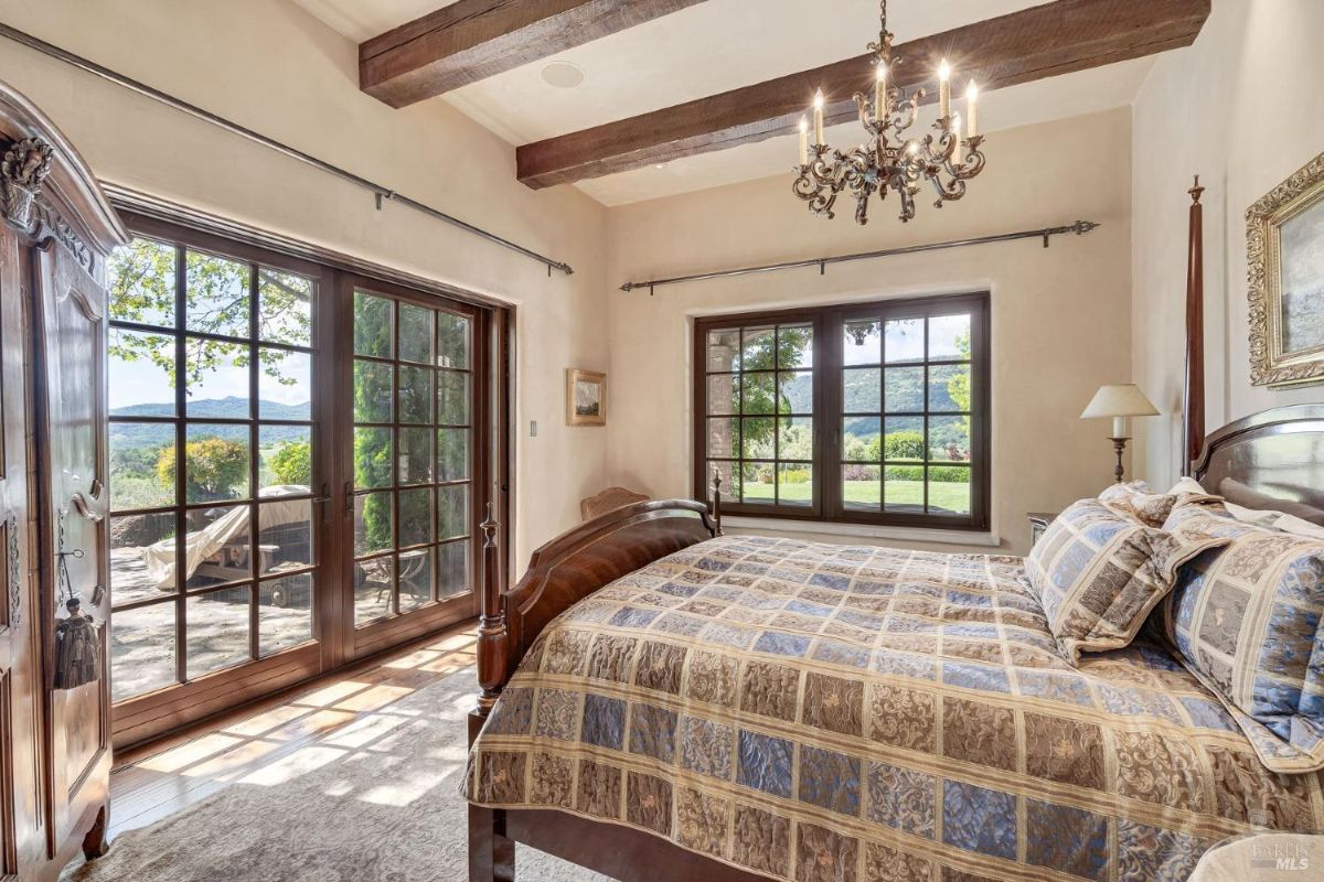 Bedroom with exposed wooden beams, large glass doors, and windows offering views of the outdoor landscape. Features a traditional wooden bed frame, patterned bedding, and a decorative chandelier.