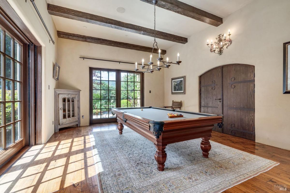 Game room featuring a pool table centered on a patterned area rug, with exposed wooden beams on the ceiling. Large glass doors provide natural light and outdoor access, complemented by rustic wooden cabinetry and a decorative chandelier.