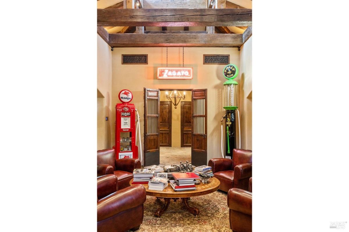 Room featuring vintage gas station memorabilia, including a red Mobilgas pump and a green Polly Gas pump. Decor includes leather armchairs arranged around a round wooden coffee table under exposed wooden beams.