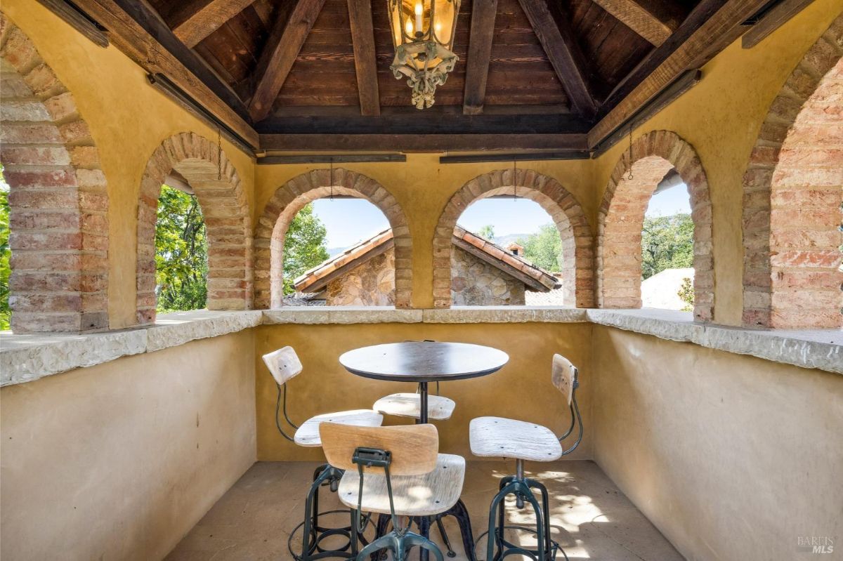 An open-air terrace with rustic brick arches, a small round table, and four chairs under a wooden ceiling.